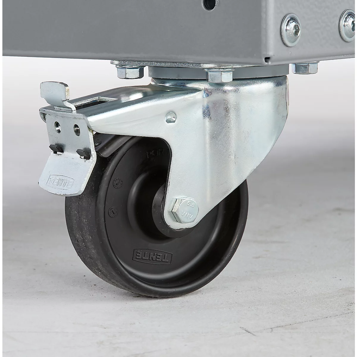 Nahaufnahme eines Rollens an einem grauen Metallgestell. Das Rad ist schwarz, das Gehäuse silbern, mit Feststellhebel.