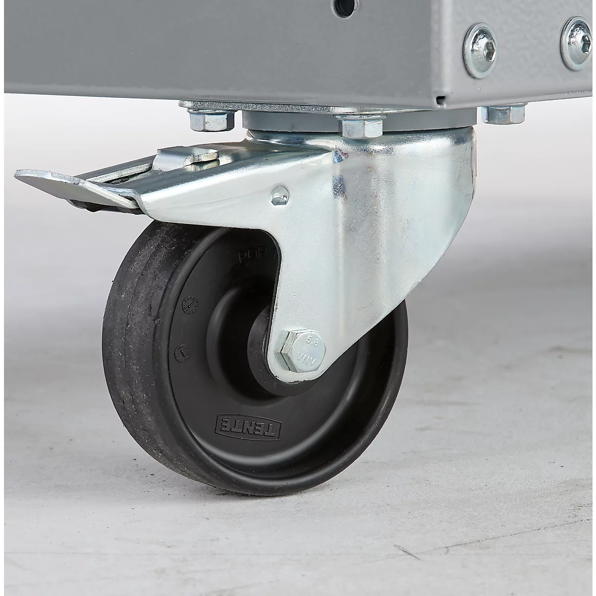 Nahaufnahme eines schwarzen Rollenrades mit Metallhalterung, verschraubt an einem grauen Metallrahmen. Grauer Boden.