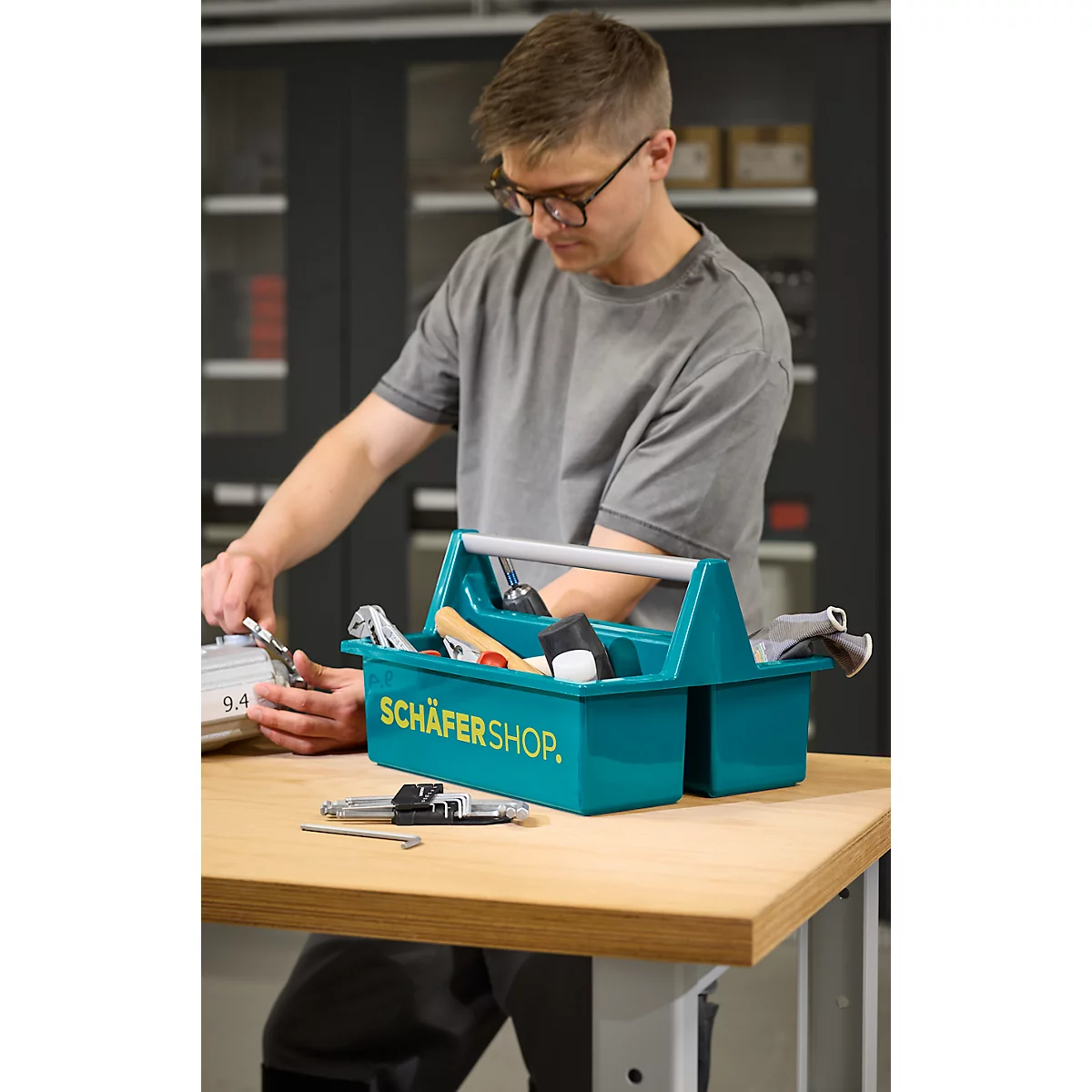 Mann mit Brille arbeitet an Tisch. Vor ihm blaue Werkzeugkiste mit Werkzeug, Aufdruck: Schäfershop.