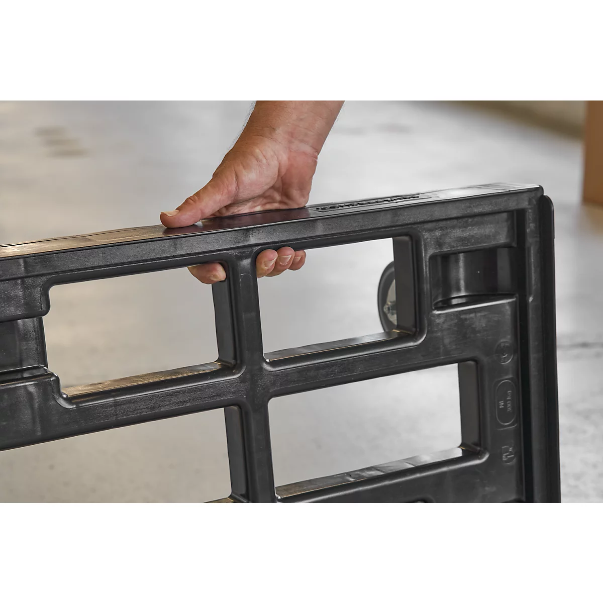 Close-up of a hand holding a black transport cart. The cart has rectangular openings and wheels.