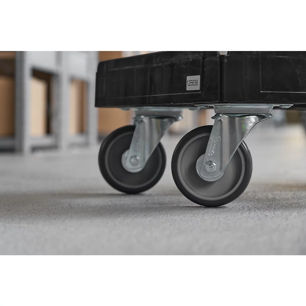 Close-up of two wheels of a black transport cart on grey floor. The background is blurred.