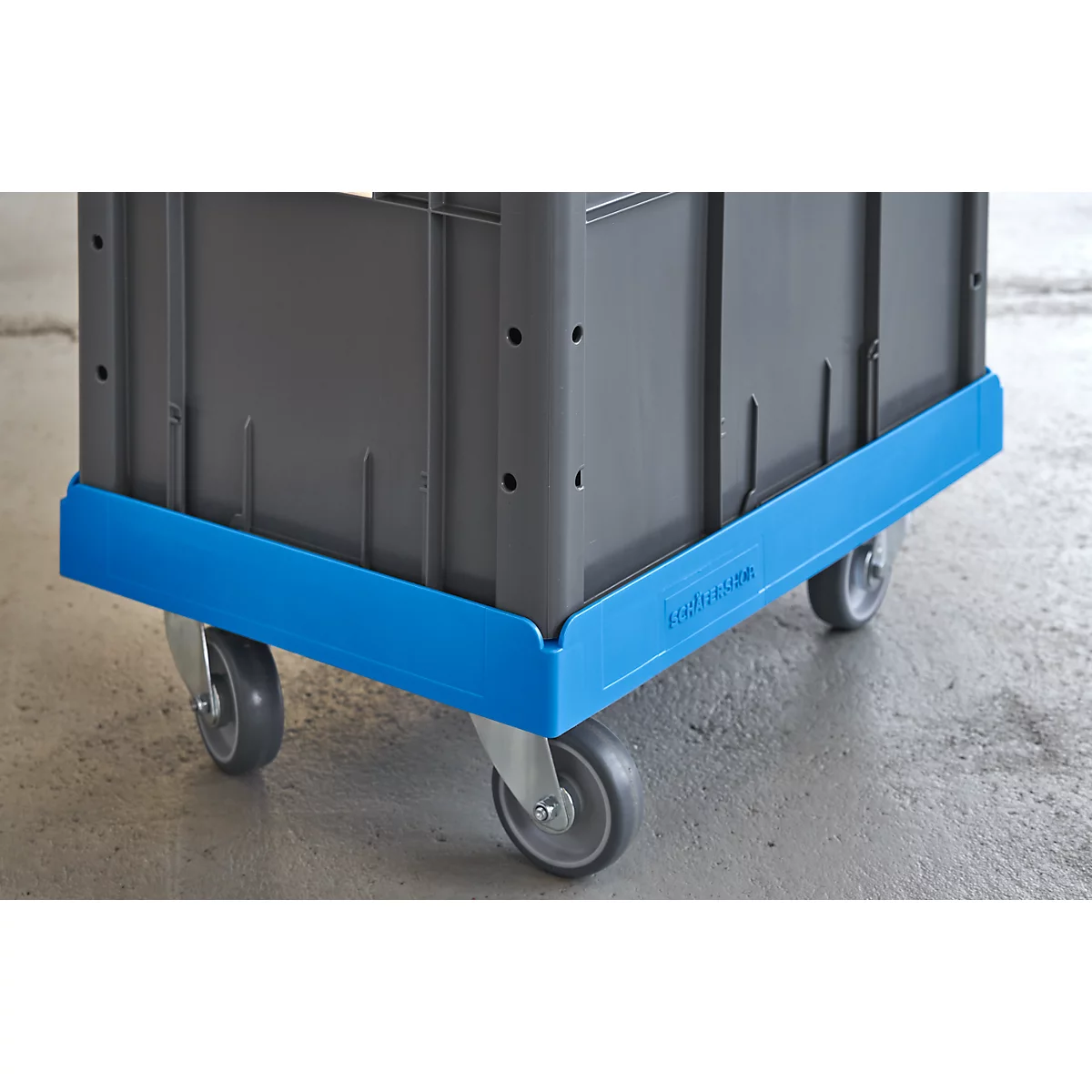 Grey transport box with blue edging and wheels on a grey floor. The box has a ribbed surface and lettering.