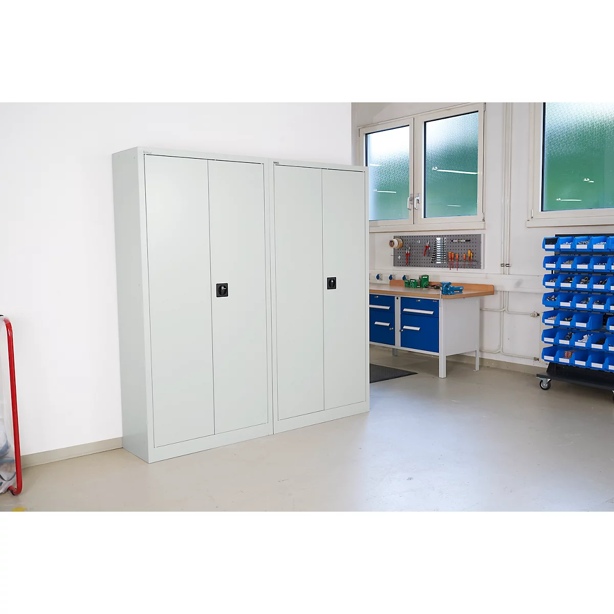 Two gray cabinets with black handles stand in front of a white wall. Tools and windows in the background.