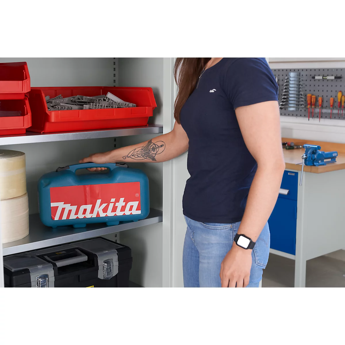 Vrouw reikt naar een Makita gereedschapskist. Makita staat in het rood op de gereedschapskist. Er zijn andere gereedschappen in de werkplaats.