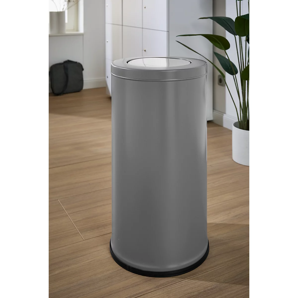 Grijze vuilnisbak in een kamer. Het deksel is licht, de basis is zwart. Op de achtergrond kasten en een plant.