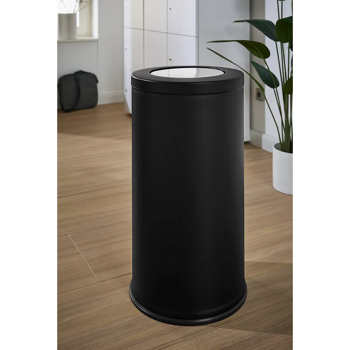Zwarte, cilindervormige prullenbak met lichtgekleurd deksel. Staat op houten vloer, achtergrond wazig.
