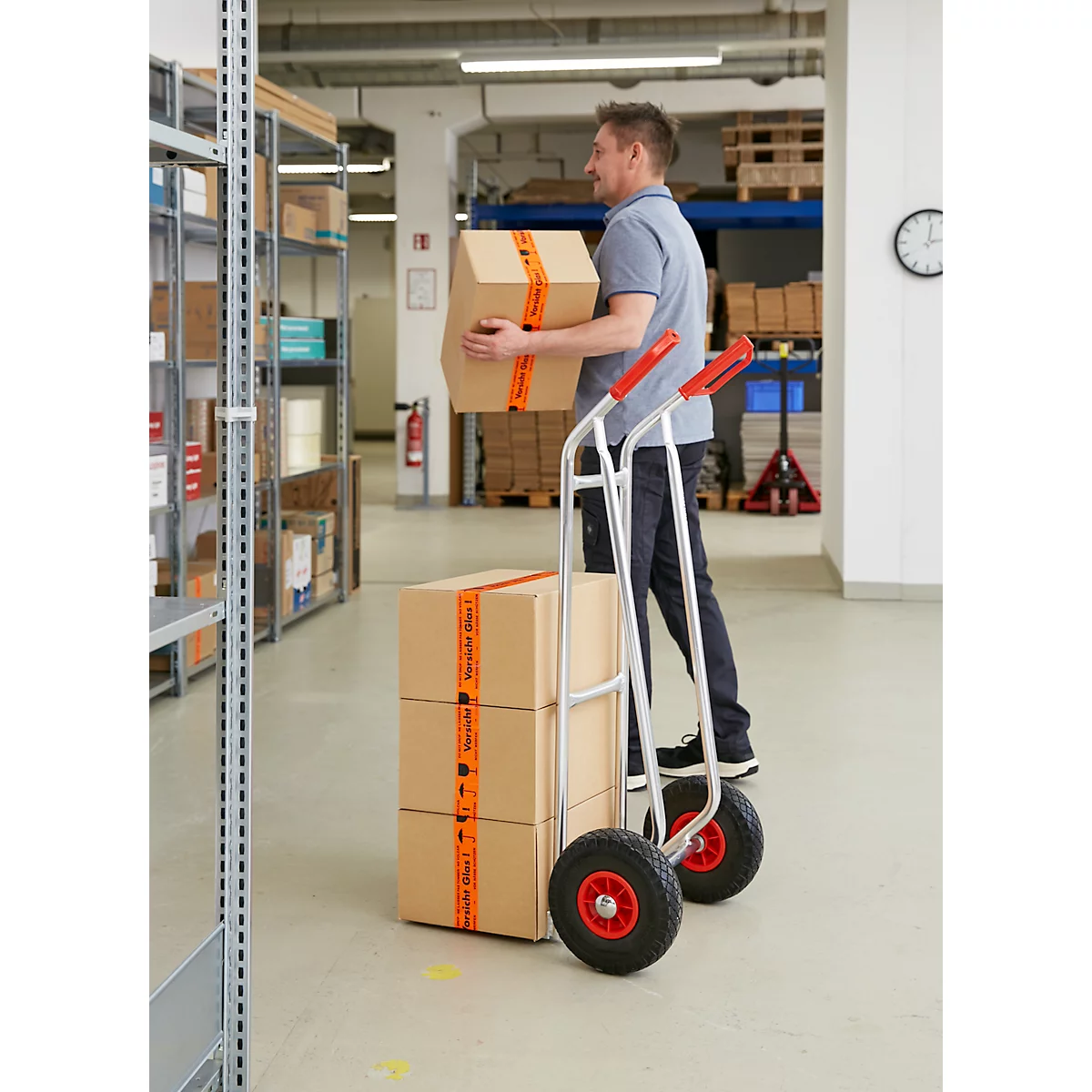 Mann transportiert Kartons mit einem Handkarren in einem Lager. Kartons sind mit orangefarbenem Klebeband versehen.