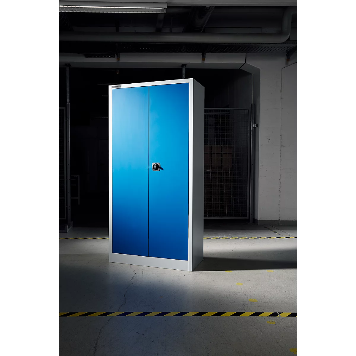 Une armoire bleue à double porte se trouve dans un entrepôt. L'armoire a un cadre argenté, une poignée noire et se trouve sur un sol en béton.