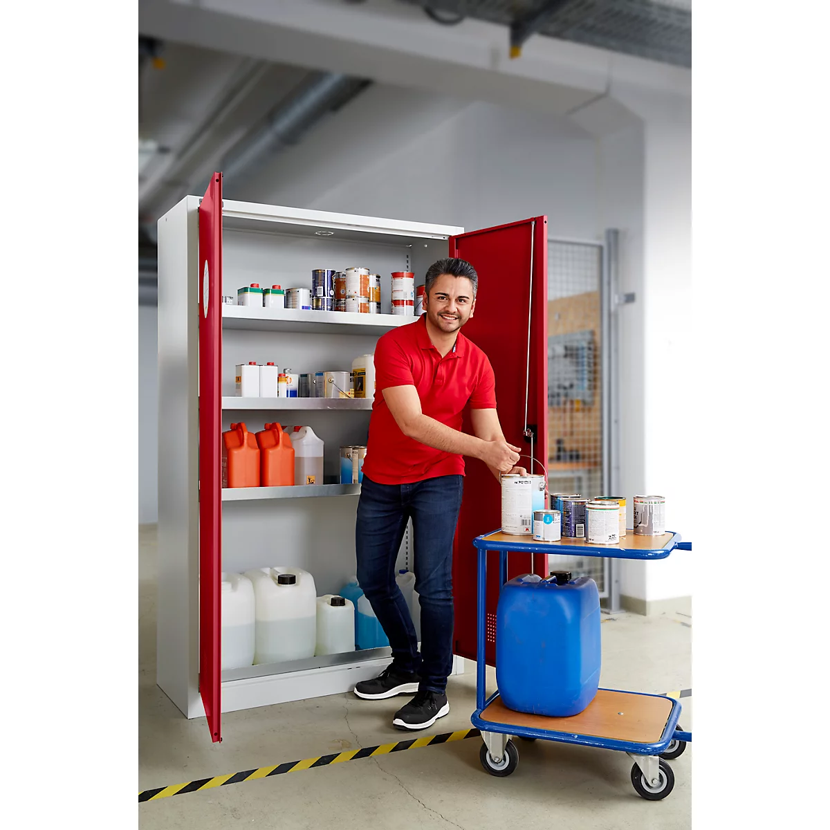 Un homme en polo rouge ouvre une armoire contenant des produits chimiques. Bidons supplémentaires sur un chariot, arrière-plan d'une pièce.