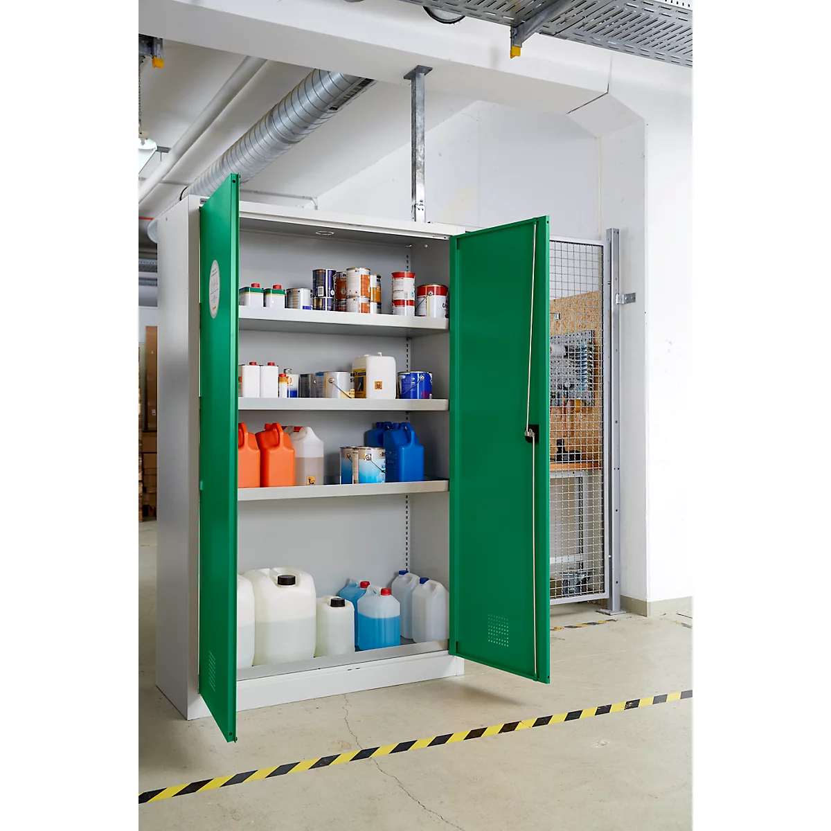 Une armoire verte ouverte contenant des produits chimiques. Divers contenants et boîtes sur les étagères. Ruban de signalisation noir et jaune sur le sol.