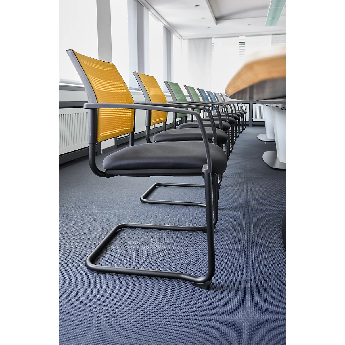 Rij zwarte vergaderstoelen met gekleurde rugleuningen. Zijaanzicht van een vergadertafel. Blauw tapijt.