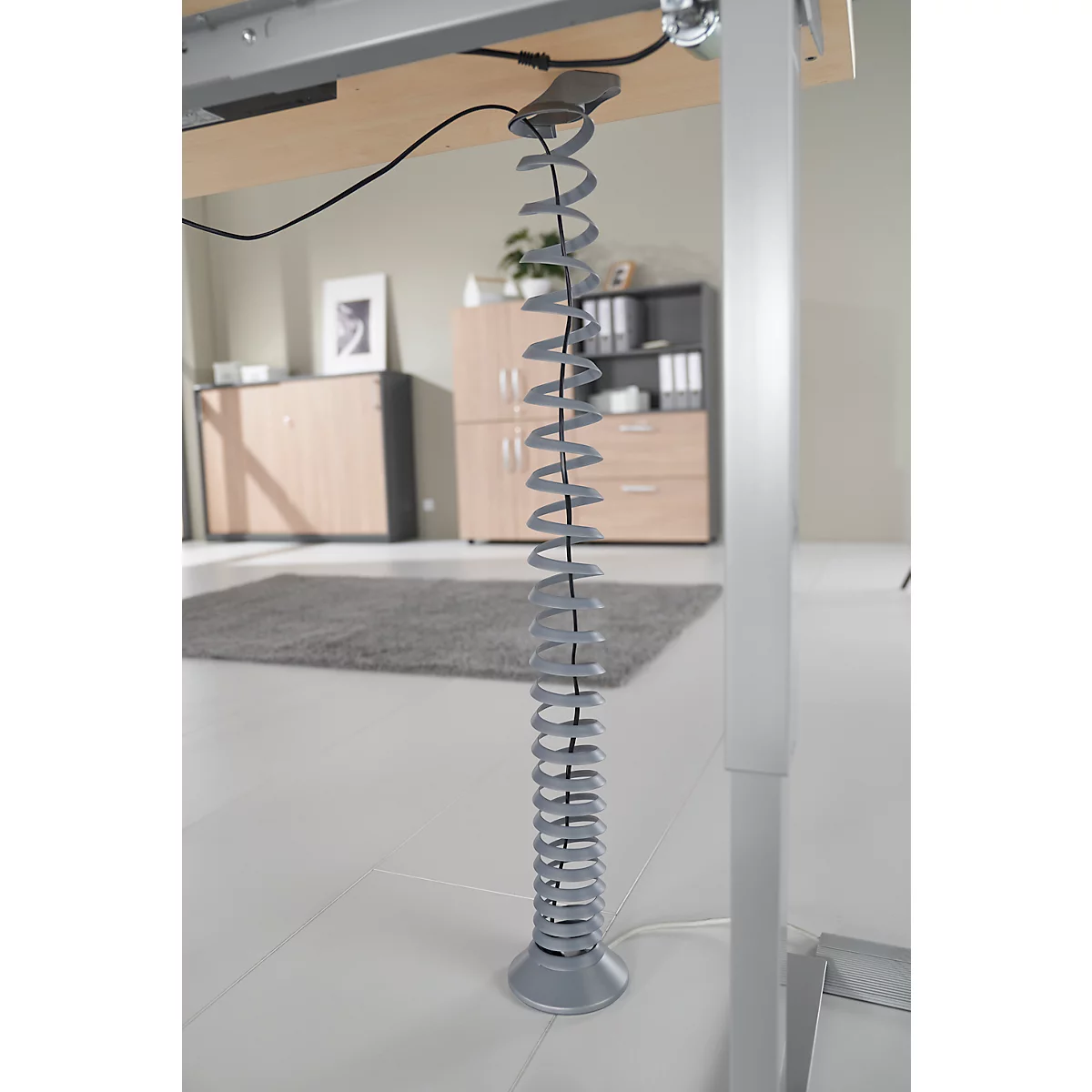 Spirale grise pour câbles sous le bureau, plan rapproché. Le bureau est clair, des armoires derrière.