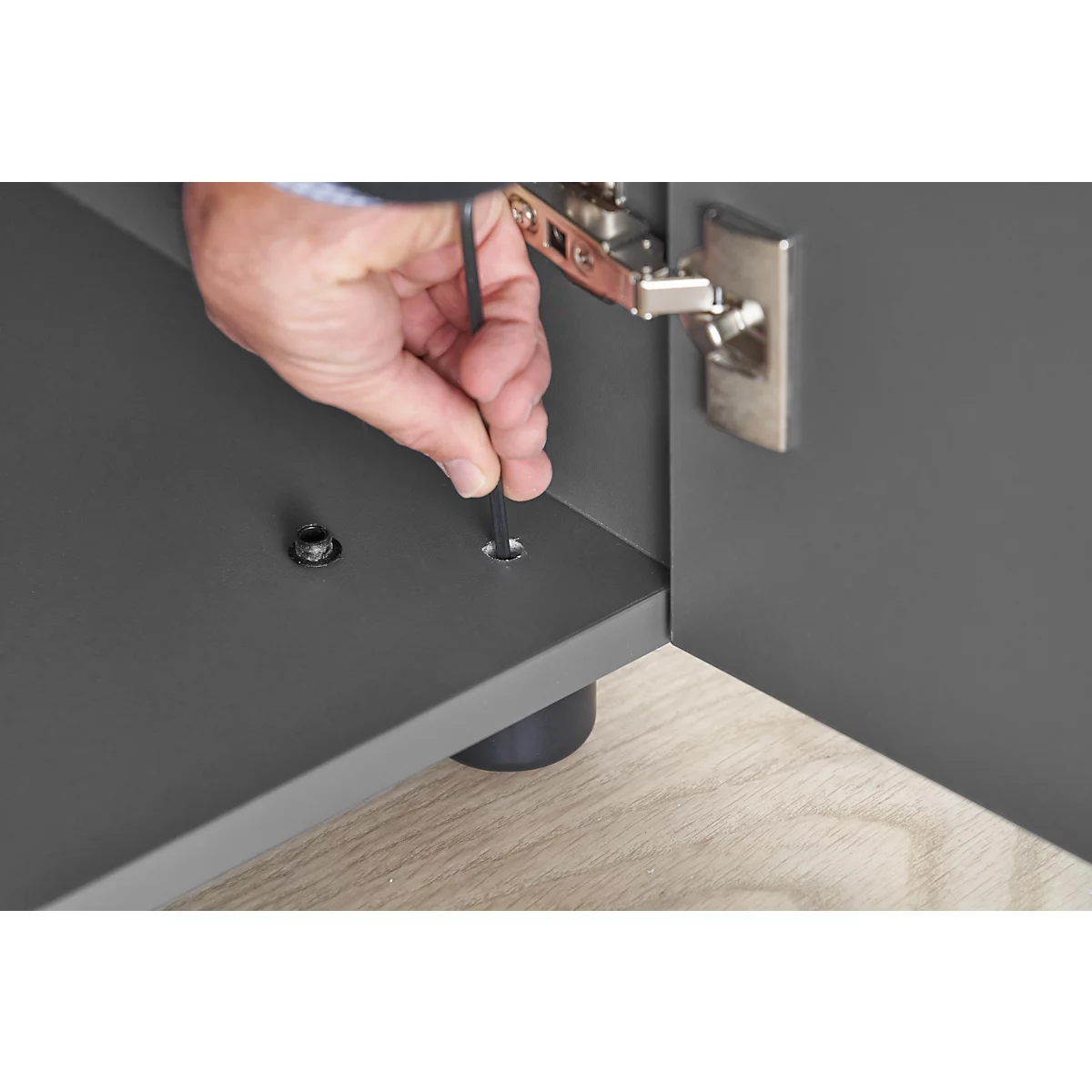Hand using an Allen key to screw a screw into a gray piece of furniture. A hinge can be seen in the background.