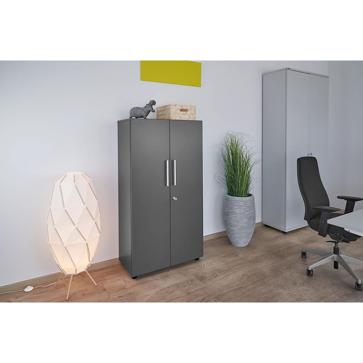 Dark grey filing cabinet in an office, next to a floor lamp and a plant.