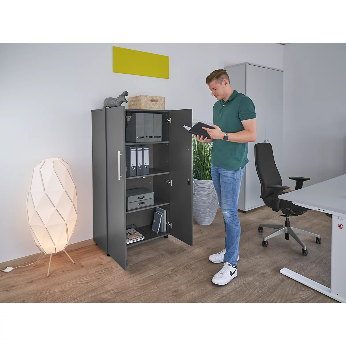 Man in office, reading notebook in front of cabinet. Gray cabinet, open. Office with lamp, table, chair.