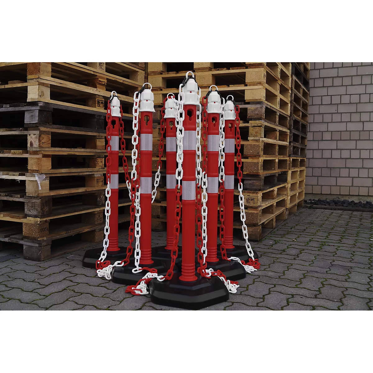 Red and white striped bollards with white chains. In the background pallets and a brick wall.