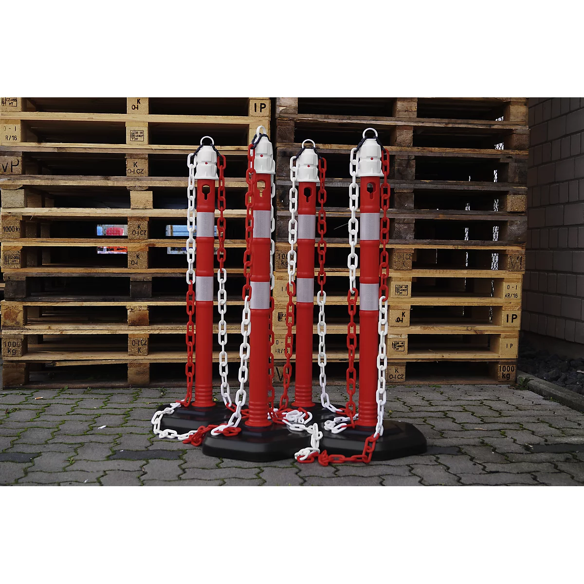 Four red and white bollards with chains in front of wooden pallets.