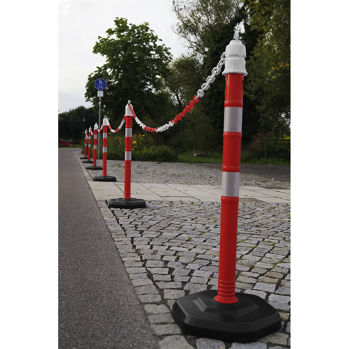 A row of red and white striped bollards with a chain on a paved path. In the background, trees and a sidewalk.