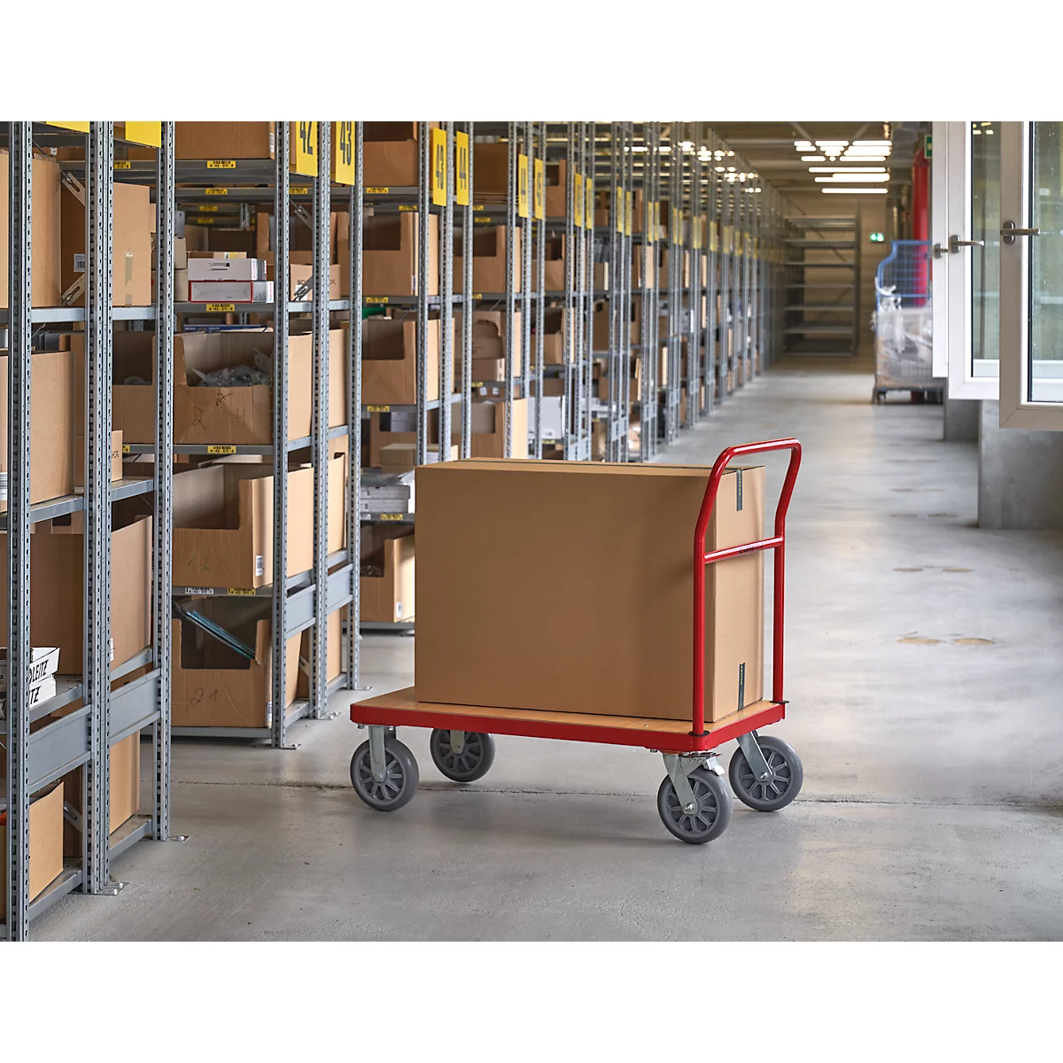 Un carton sur un chariot rouge dans un entrepôt. Rayonnages de boîtes à gauche, allées en arrière-plan.