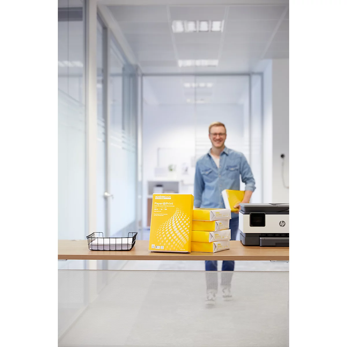 A man stands in the office. Stacks of paper and a printer are on a table.