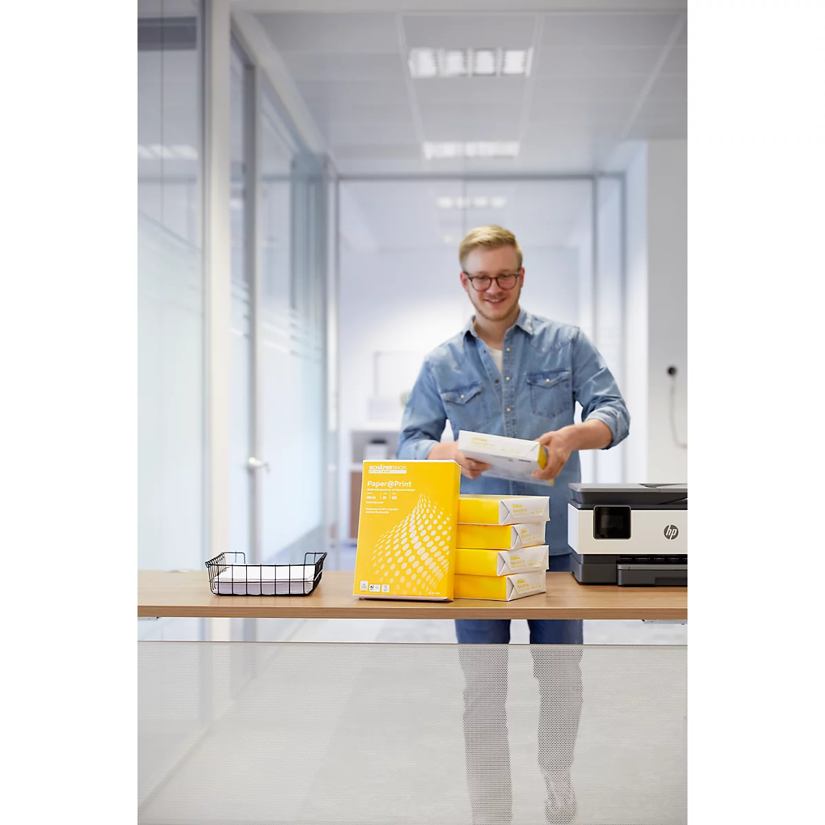 A man with glasses stands smiling at a table with a printer and paper, holding paper in his hand. Yellow paper stacks next to it.