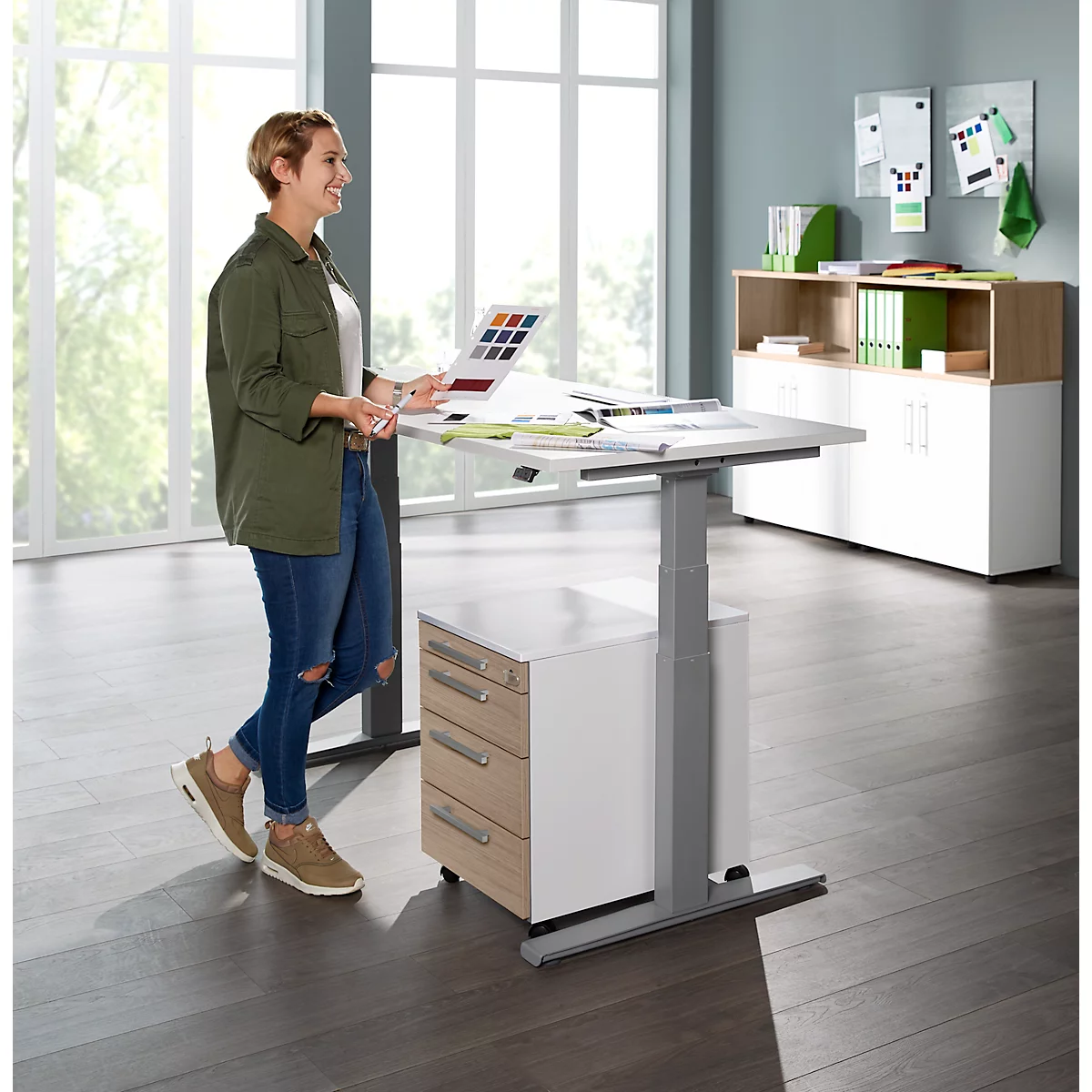 A woman stands at a standing desk, looking at color samples. She wears jeans and a jacket. The desk is light, the background gray.