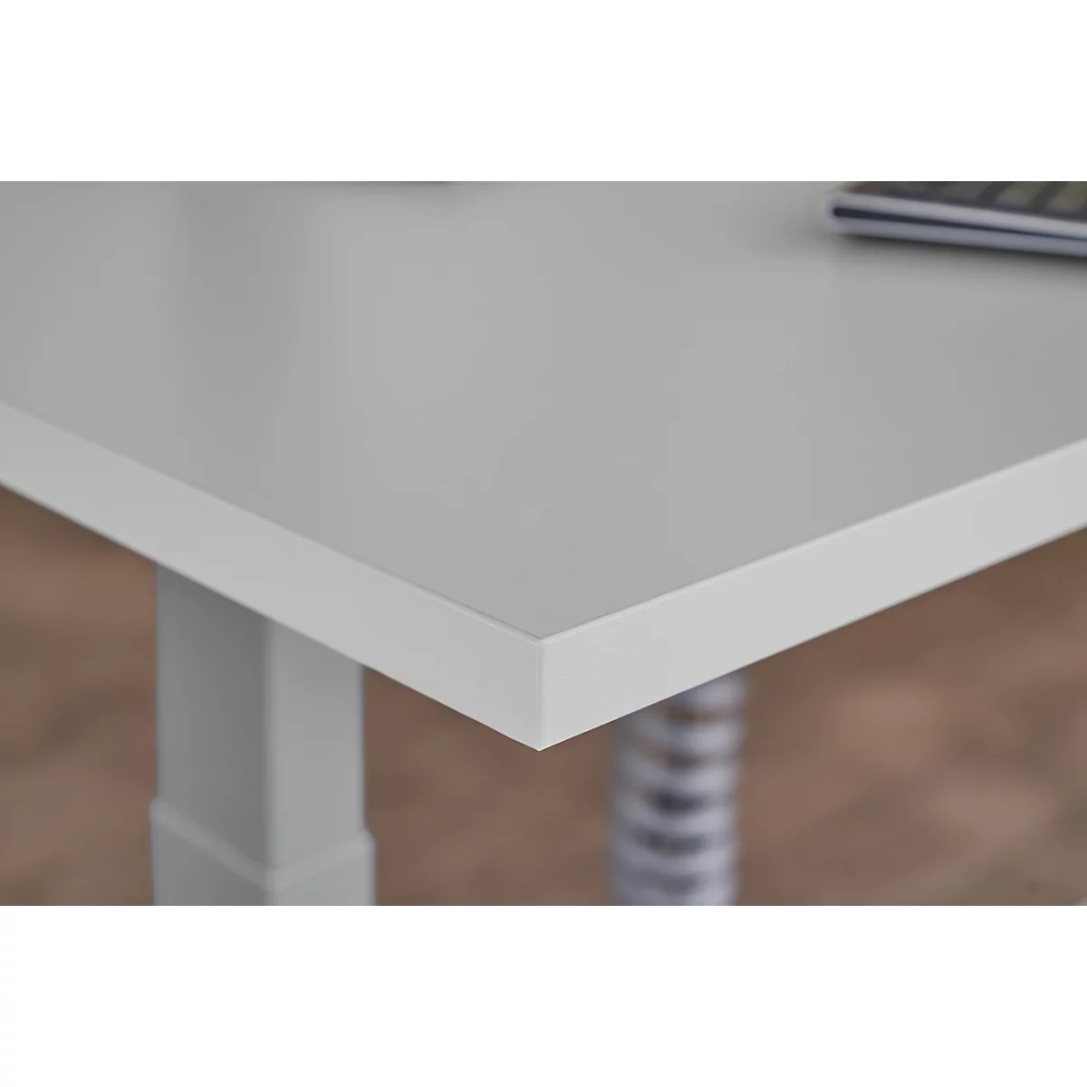 Close-up of a light grey table top with white edge, in the corner area. Underneath is a table leg.