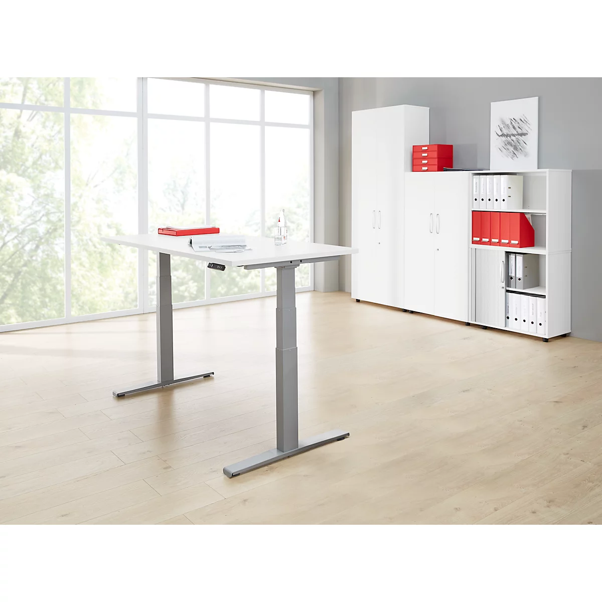 A white, height-adjustable desk stands in a bright office in front of a window and white cabinets.