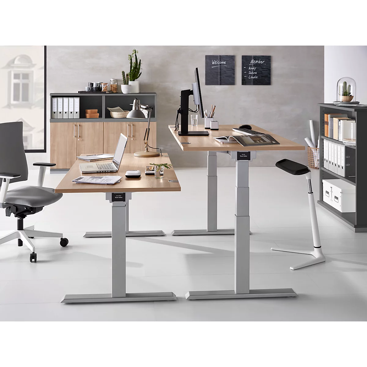 Office with two height-adjustable desks, office chairs, and shelving. Laptops and accessories are on the tables.