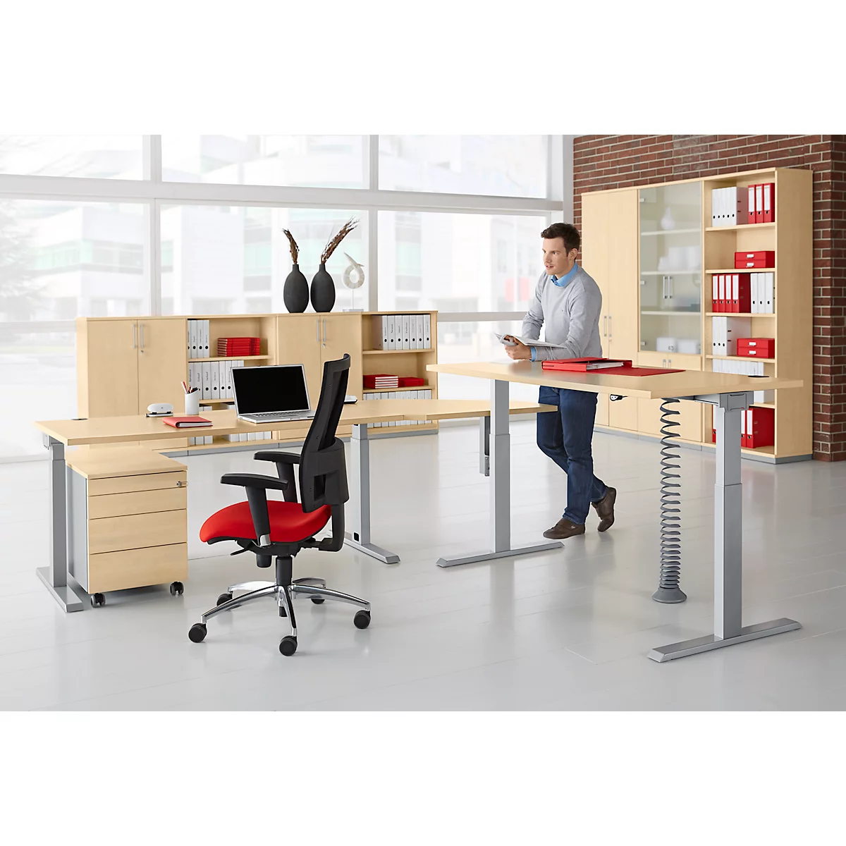 A man stands near a height-adjustable desk. Office equipment: laptop, chair, cabinets. Decoration: vases.