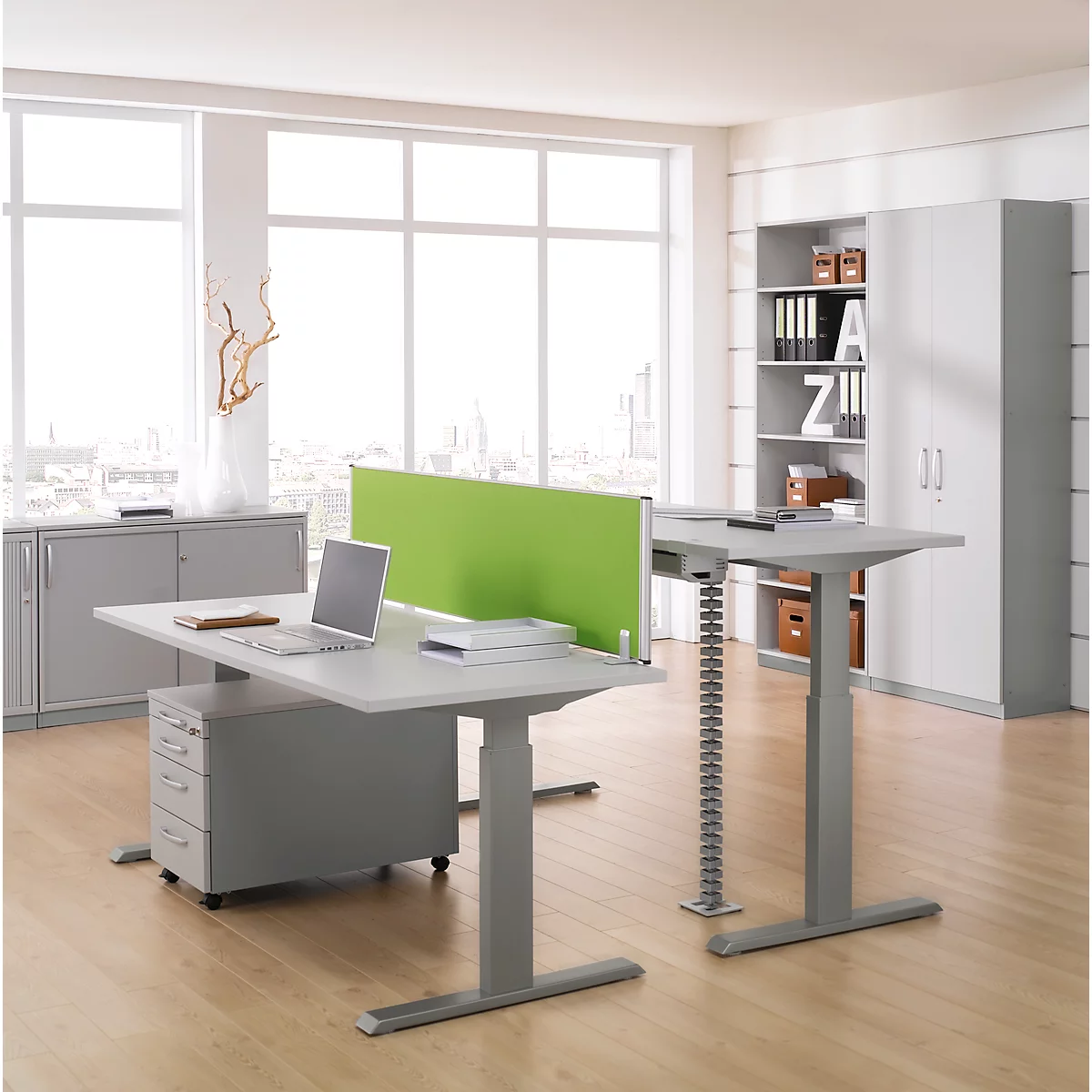 Bright office space with height-adjustable desks, laptop, file cabinet, green partition, and a view of a city.