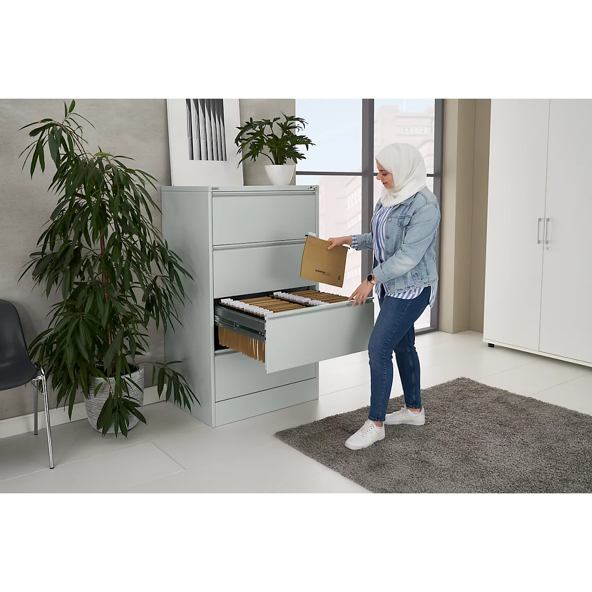 A woman in jeans and a denim jacket places a file in a gray filing cabinet. There is a houseplant next to the cabinet.