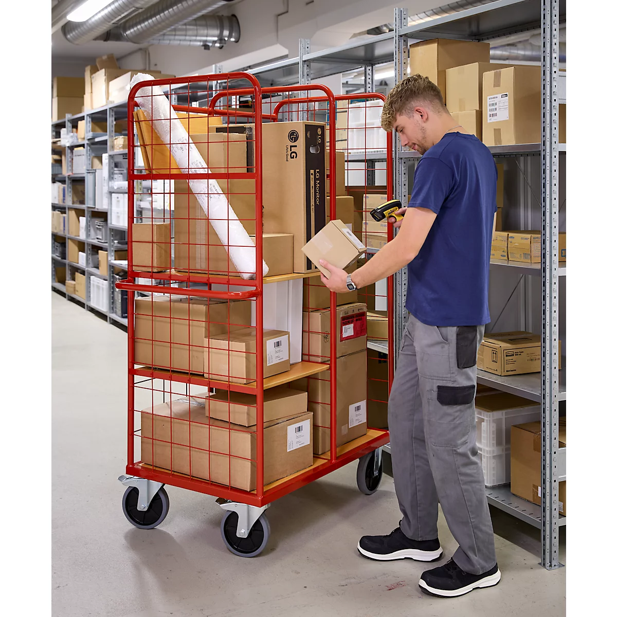 Ein Mann in Arbeitskleidung scannt Pakete in einem roten Transportwagen im Lager. Regale mit Kartons im Hintergrund.