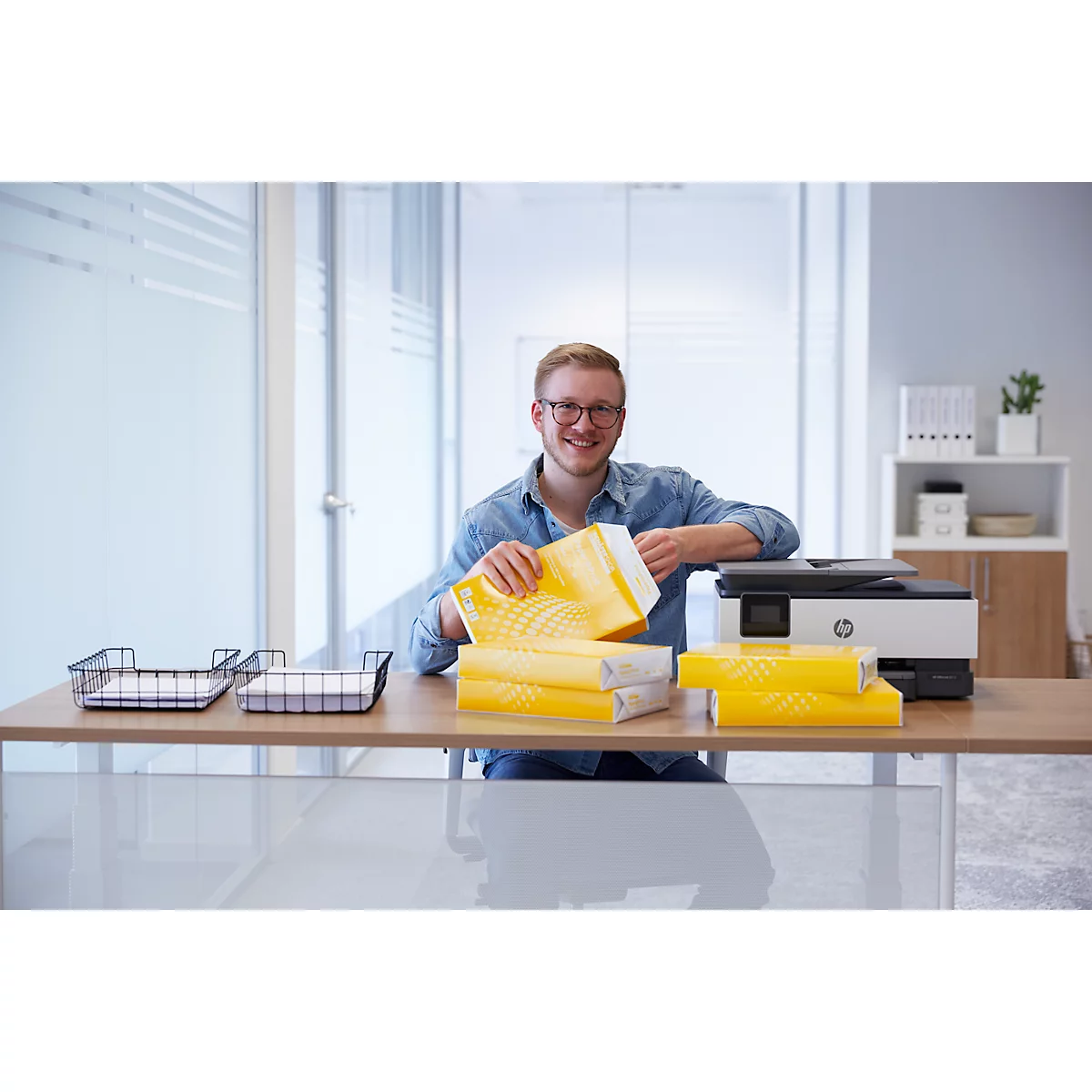 Ein Mann mit Brille öffnet Papierverpackung am Schreibtisch. Auf dem Tisch steht ein Drucker.
