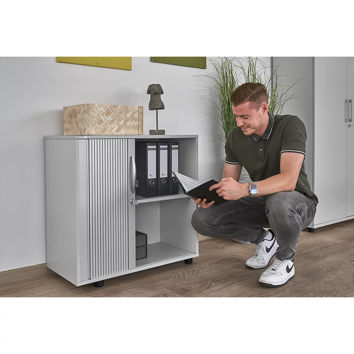 Man squats by a gray cabinet, reading a book. The cabinet has a roller shutter. File folders sit on the shelf.