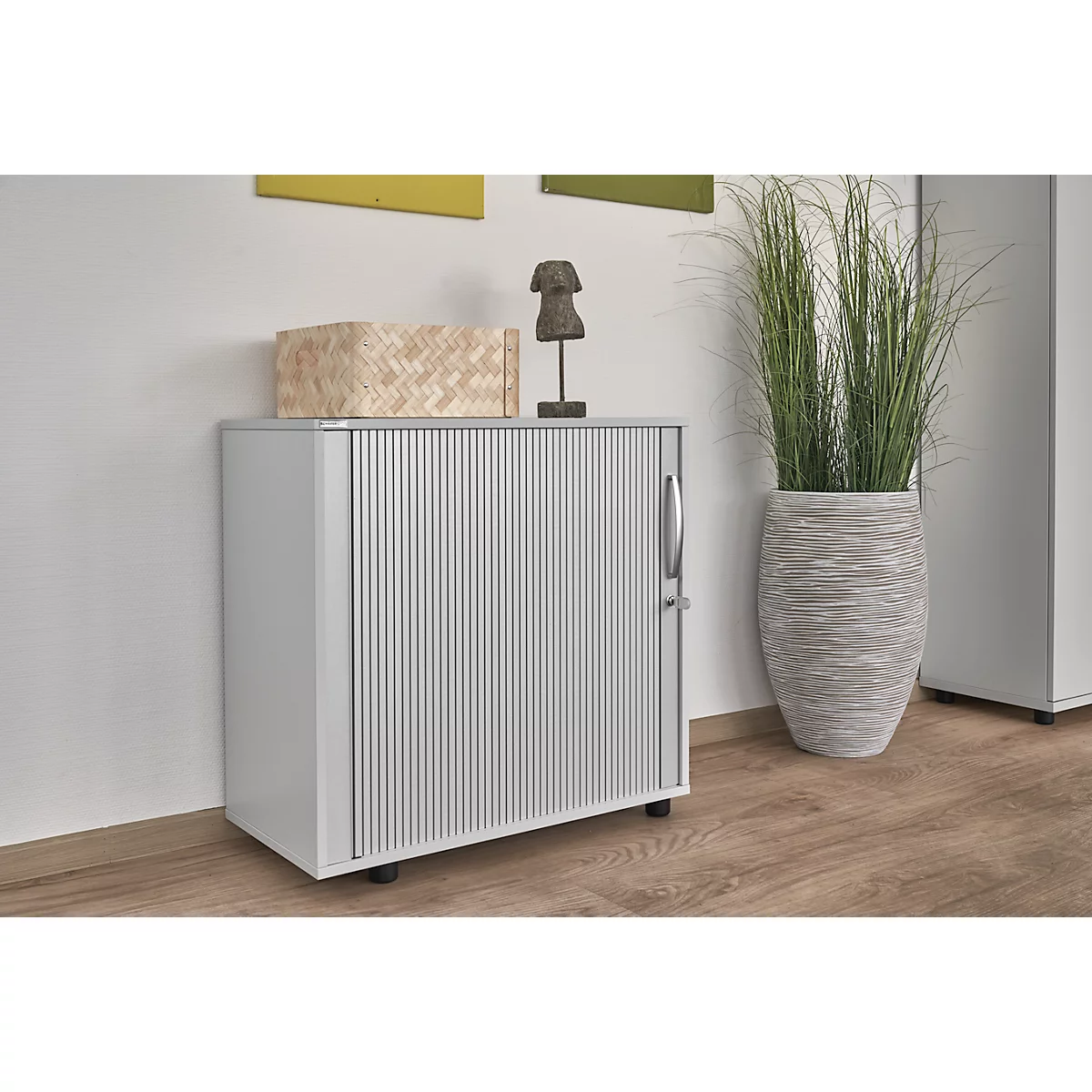 Grey cabinet with ribbed front and decor on top. To the right, a tall vase with ornamental grass.