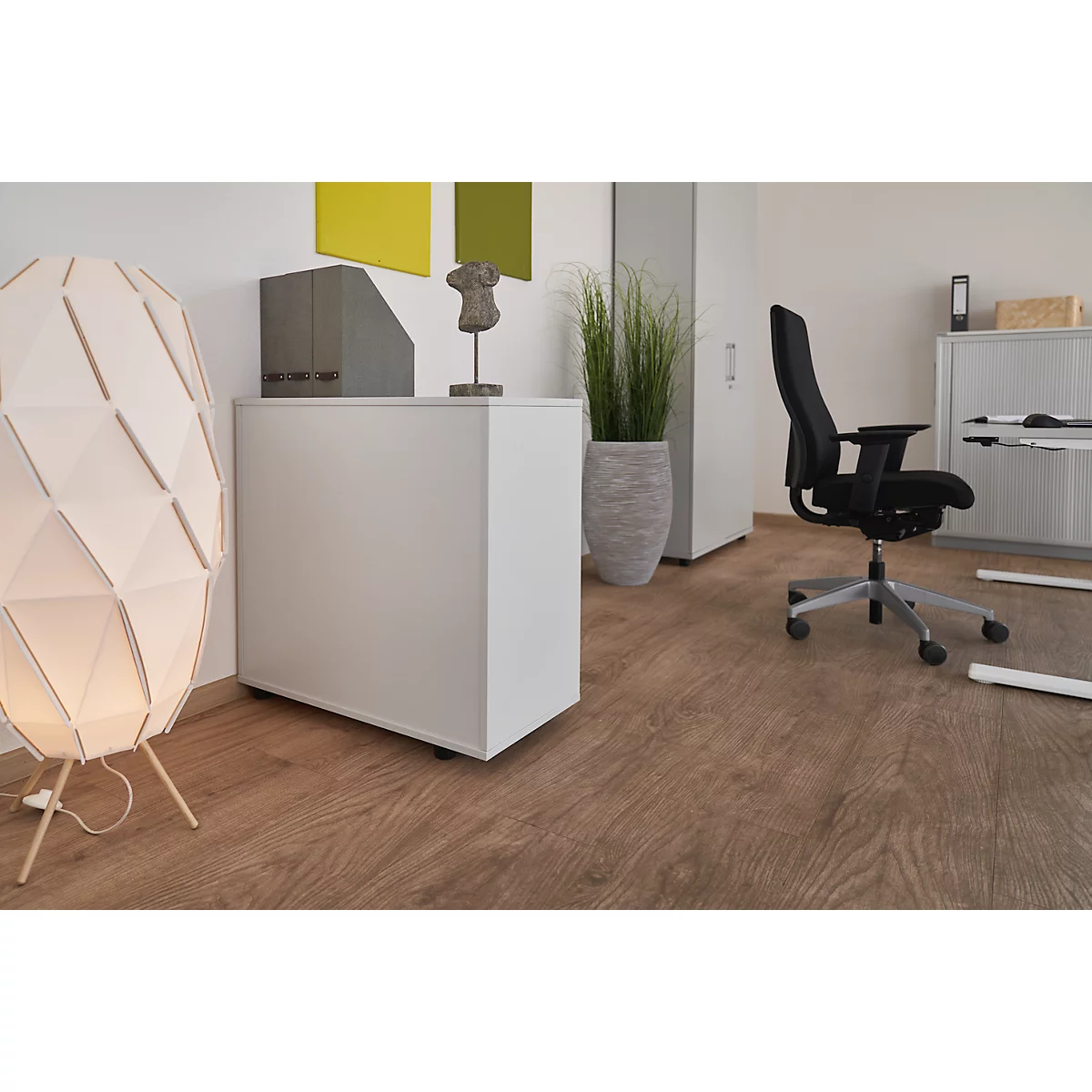 Office with standing desk, office chair, lamp, plant. Wooden floor, white walls, bright furnishings.