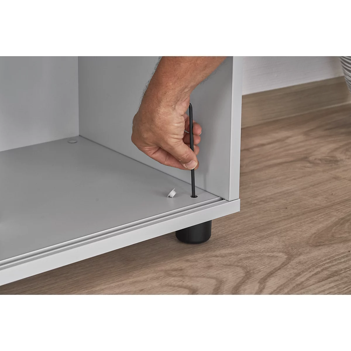 Hand with Allen key inserts screw into furniture. Furniture stands on black leg. Wooden floor.