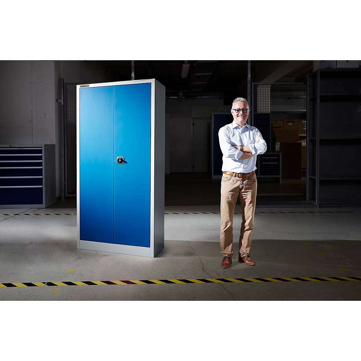 Man in magazijn voor blauwe kast. Hij draagt overhemd, broek, bril en gekruiste armen.
