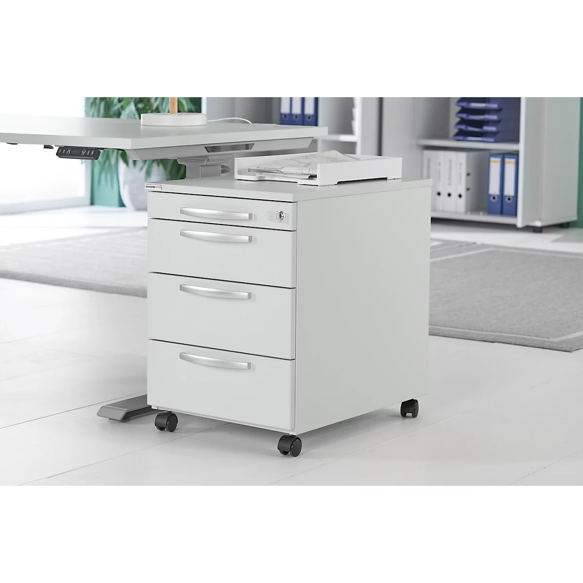 A grey, three-drawer cabinet with castors stands next to a height-adjustable desk.
