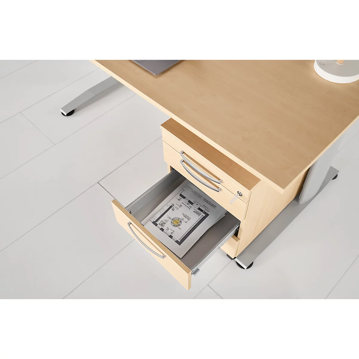 Office desk with open drawer. There is a document in it. The desk has a wood look and silver legs.