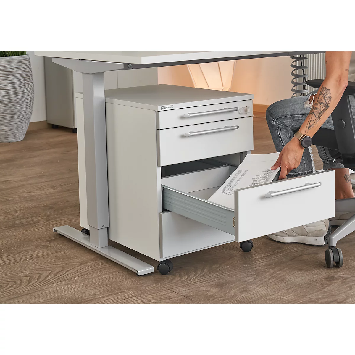 Person opening a drawer of a white mobile pedestal under a desk. Papers are inside.