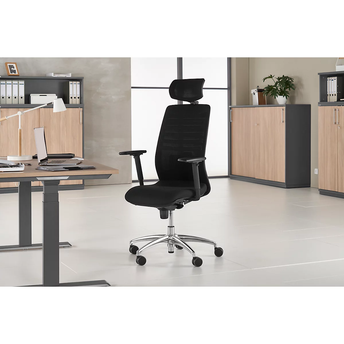 Black office chair with headrest, in front of a bright background. Desk, cabinet and window in the background.