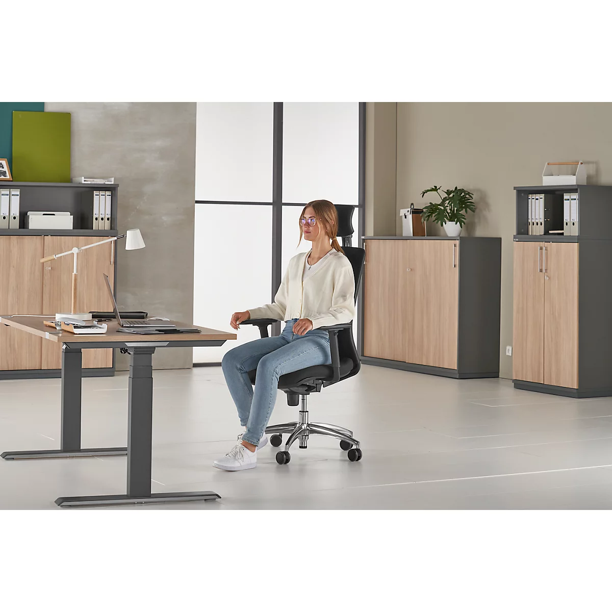 Woman sits at a height-adjustable desk, working on a laptop. The room looks bright and modern.
