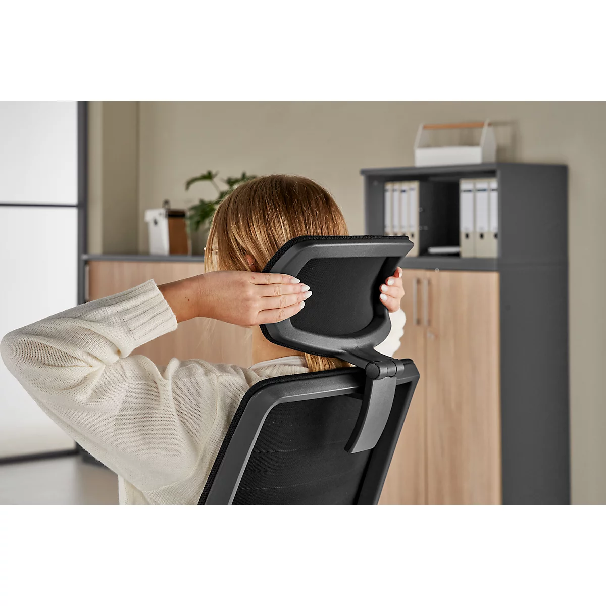 A woman rests her head against an office chair and touches her neck.