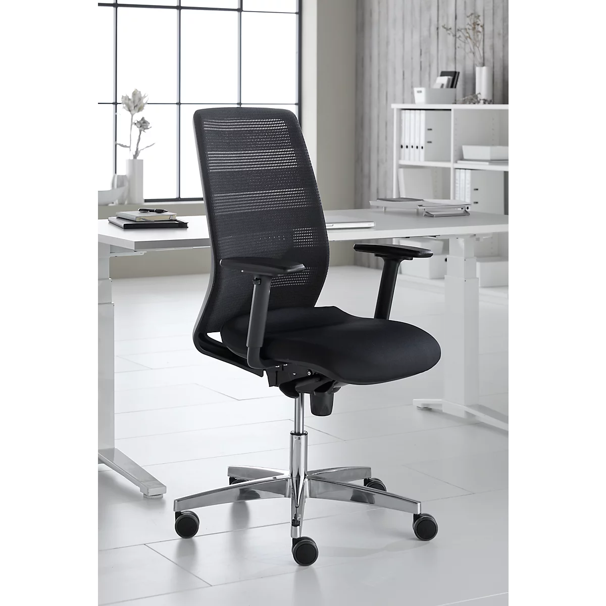 Black office chair, mesh back, armrests, chrome base. Stands in front of a window and desk.