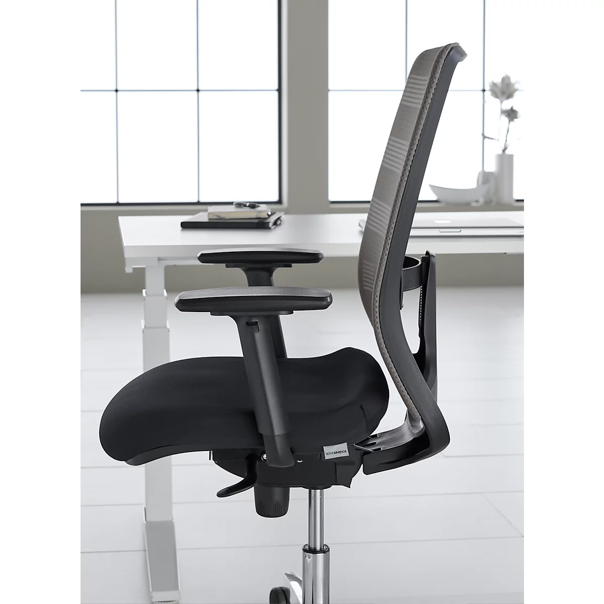 Black office chair in profile against a light background. The chair has a mesh back and adjustable armrests.
