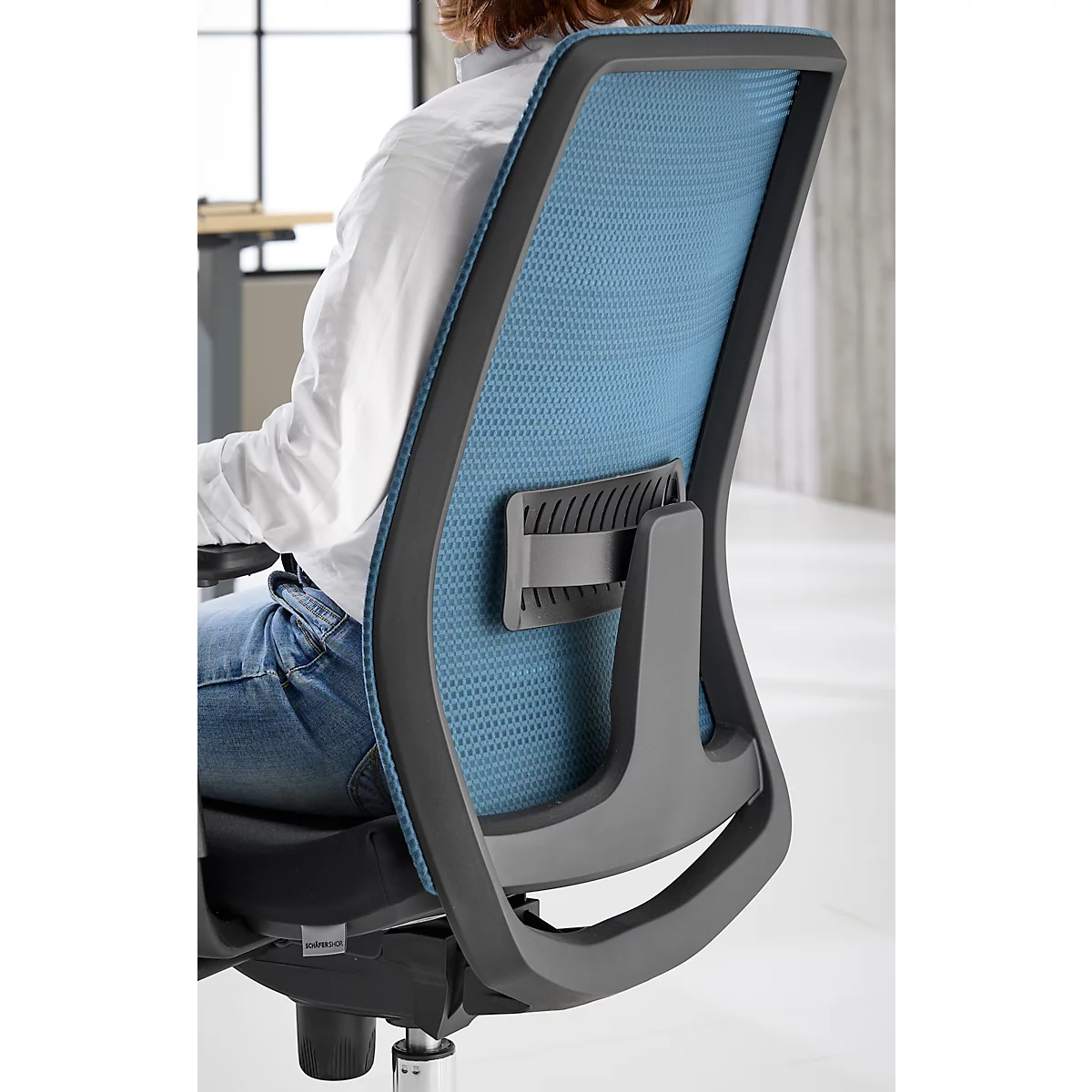 Back of an office chair. Blue mesh structure with gray frame and bracket, in front of person in white blouse.