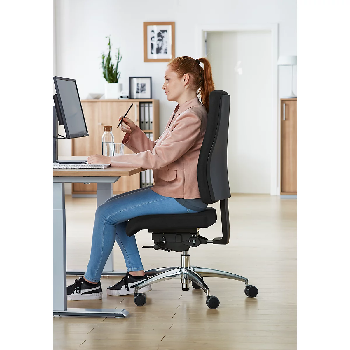 A red-haired woman in a pink blazer is sitting at a desk, using an office chair. She is working on the computer and holding a pen.