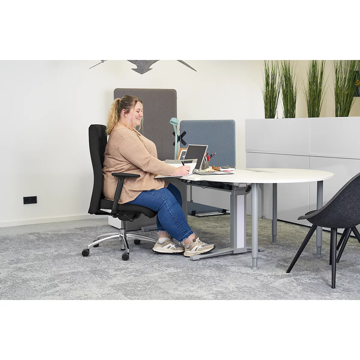 A smiling woman sits at a desk, writing something.